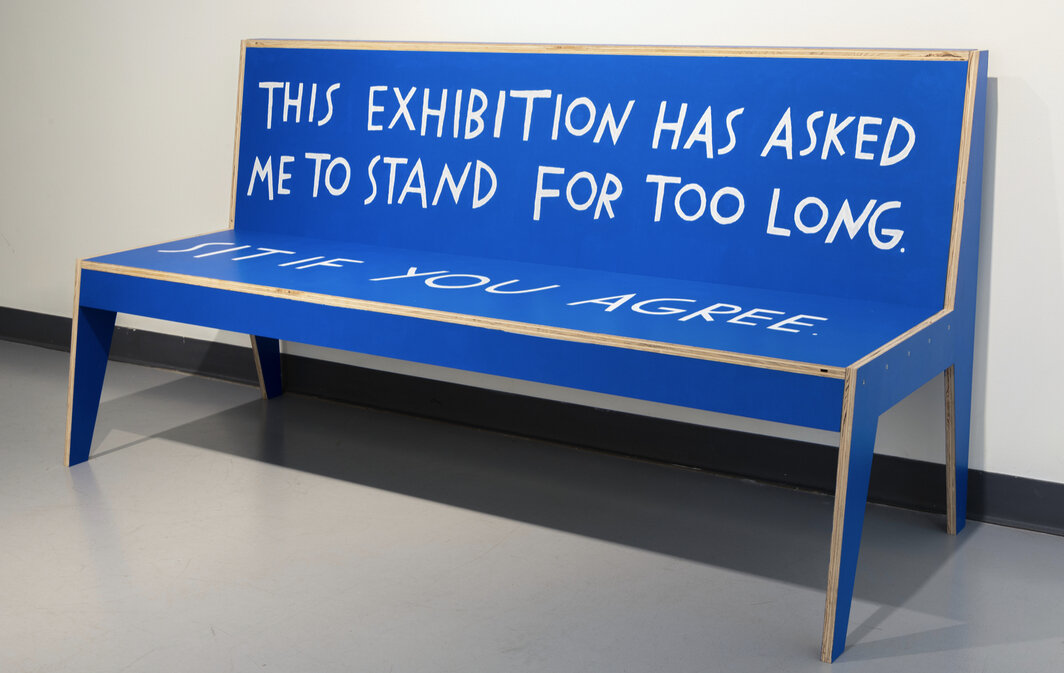 blue bench made of plywood, which is exposed at the joins. On the back it has text saying “This exhibition has asked me to stand for too long” and on the seat it says “Sit if you agree”