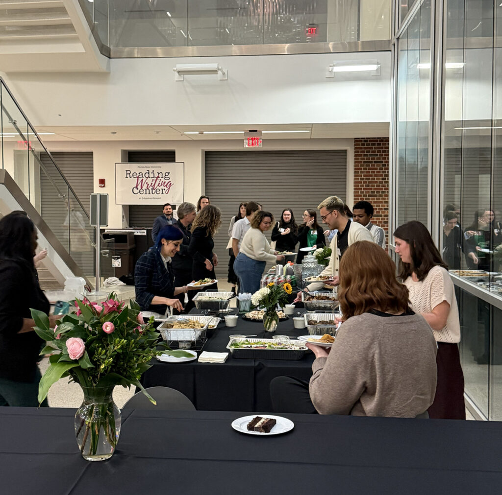 there are many people standing around a table with plates of food on it