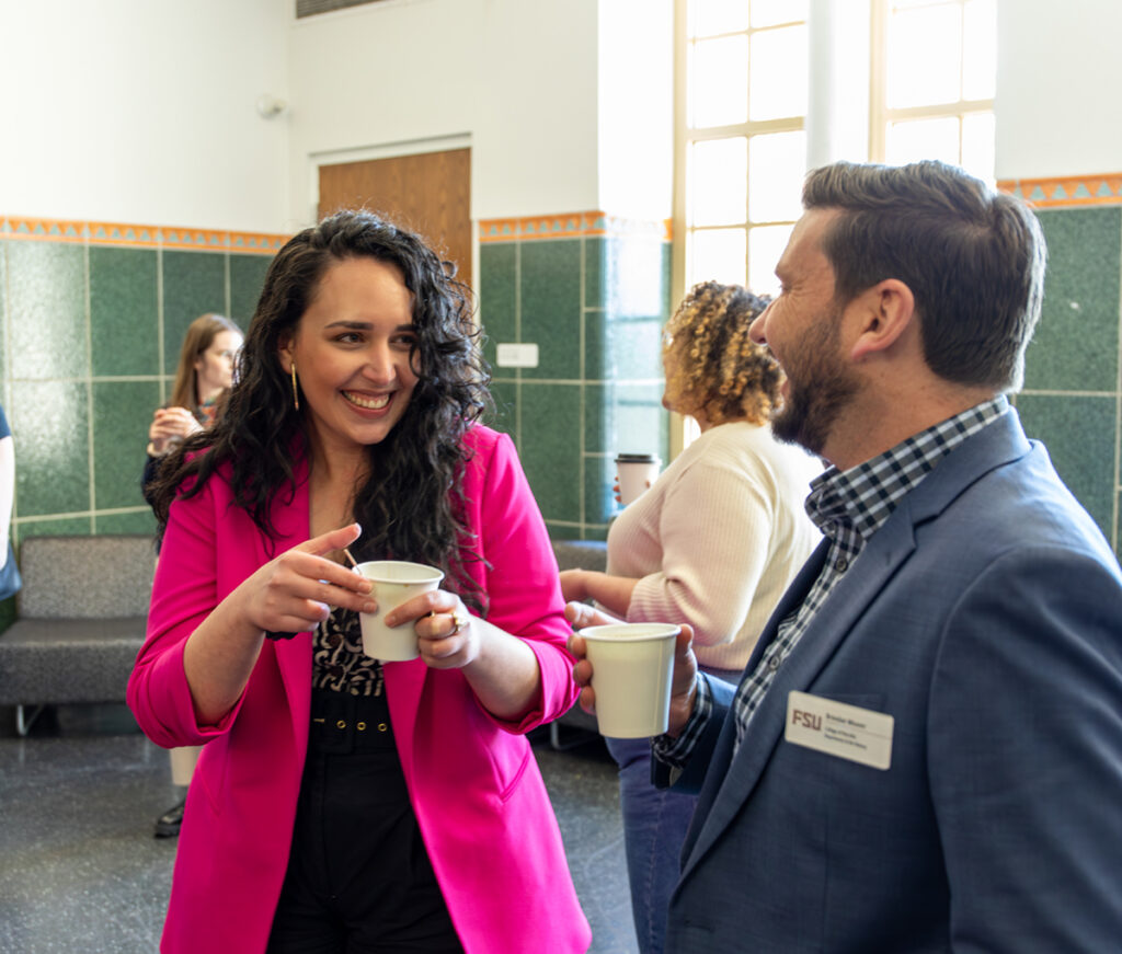 smiling woman in pink blazer holding a cup of coffee talking to a man in a pink blazer